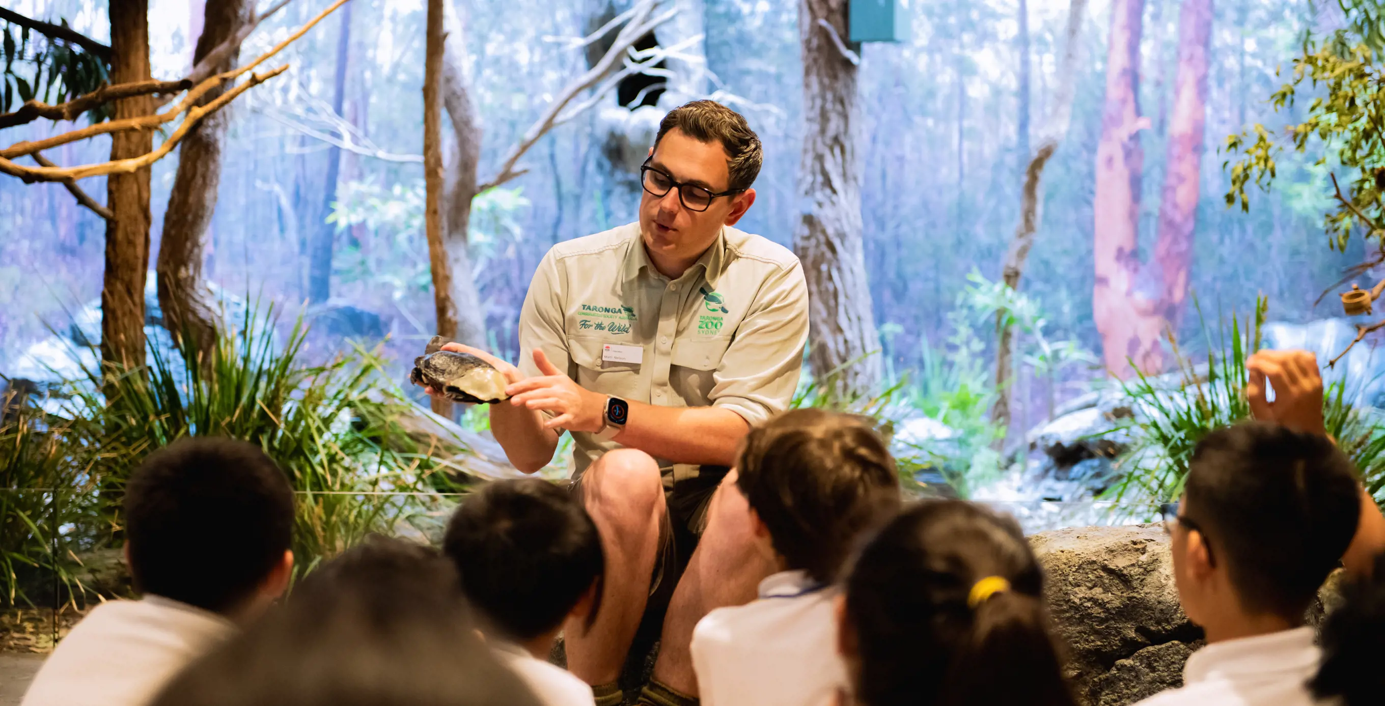 School students seated on the floor during a teacher‑led lesson in an immersive habitat classroom.