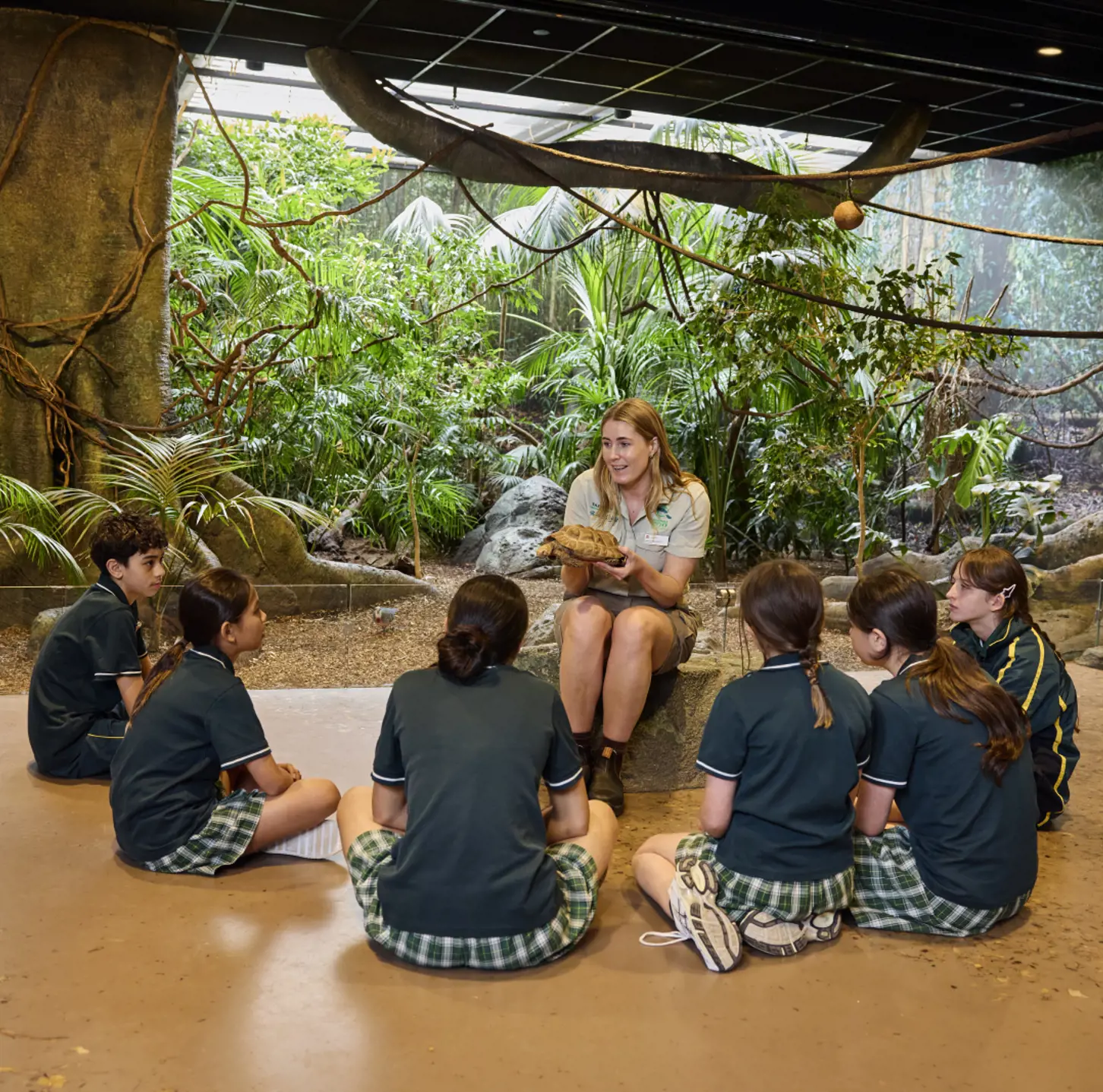 Children in a classroom listening to a teacher with greenery in background.
